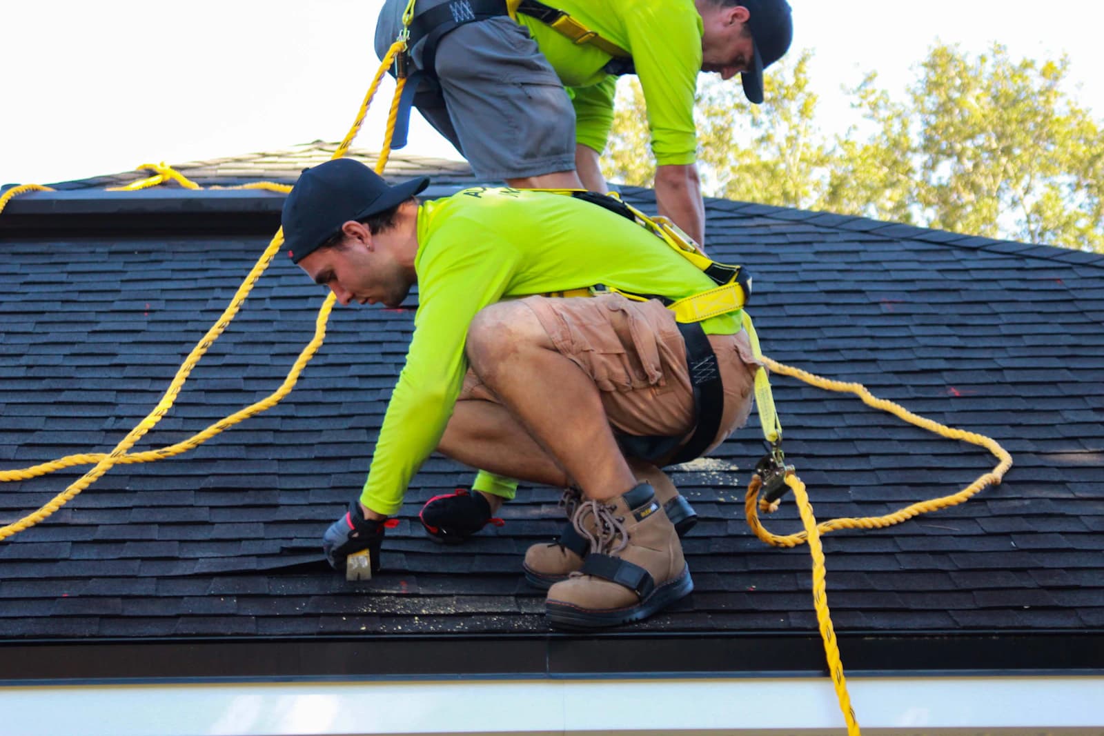 Two roofers in safety harnesses installing shingles on a residential roof